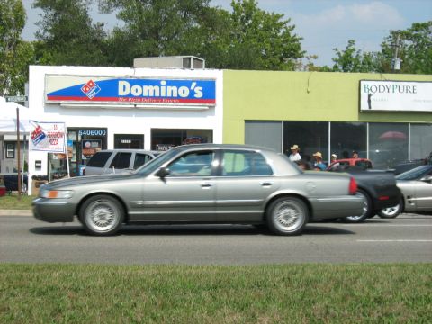1988 Mercury Grand Marquis - A silver Mercury Grand Marquis sedan is seen driving on a street in front of commercial buildings.