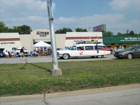 1988 Mercury Grand Marquis - A white Ecto-1 car, resembling the Ghostbusters vehicle, is seen at an outdoor event with other cars and people.