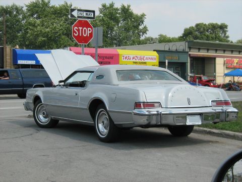 1988 Mercury Grand Marquis - A silver 1977 Lincoln Continental Mark V coupe is parked with its hood up, near a stop sign and businesses.