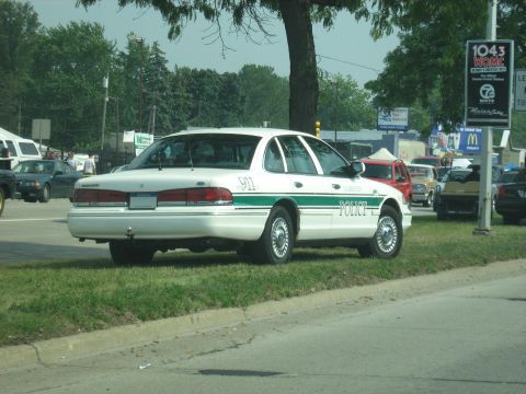 1988 Mercury Grand Marquis - A police car is parked on the grass next to a road, with other vehicles and tents in the background.