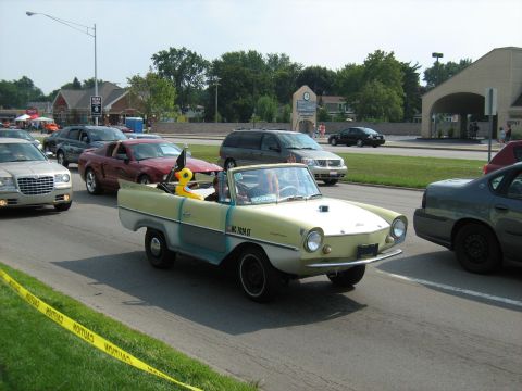 1988 Mercury Grand Marquis - An amphibious car drives in a parade, accompanied by other vehicles and a caution tape barrier.