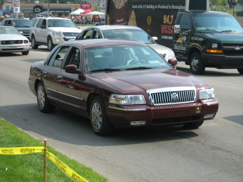 1988 Mercury Grand Marquis - A maroon sedan drives down a street with other vehicles and tents in the background.
