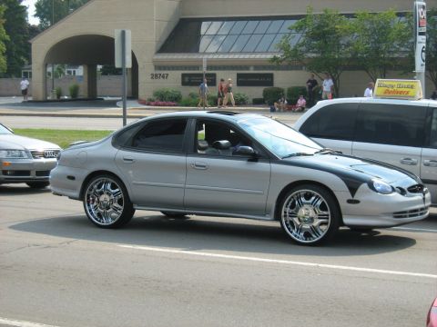 1988 Mercury Grand Marquis - A silver sedan with custom rims is parked on a street next to a white van.