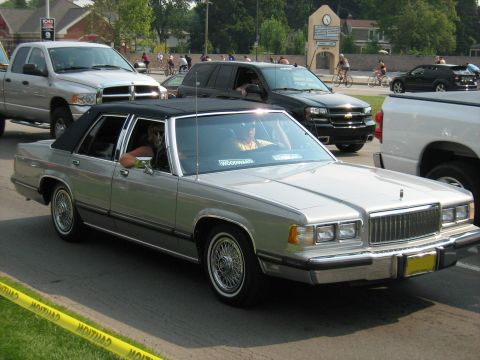 1988 Mercury Grand Marquis - A silver 1988 Mercury Grand Marquis with a black vinyl roof cruises down the street, seen from a 3q-front perspective.
