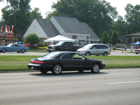1988 Mercury Grand Marquis - A black coupe drives by a street scene with a BBQ stand and other vehicles.