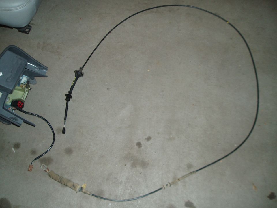 Removed shift cable coiled on a concrete floor beside a floor shifter console assembly