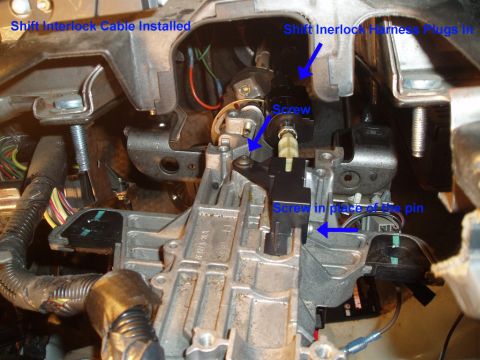 2003 Ford Crown Victoria - Close-up of a disassembled steering column showing the shift interlock cable and harness area, with blue text annotations pointing to the i…
