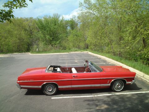 1967 Ford Galaxie - A red 1967 Ford Galaxie convertible is parked in a lot with trees in the background.