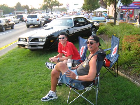 1988 Mercury Grand Marquis - Two men relax on chairs next to a black Pontiac Firebird Trans Am at an outdoor car event.