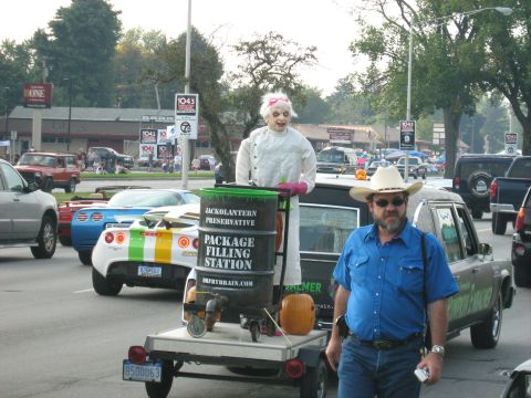1988 Mercury Grand Marquis - A man stands by a hearse decorated with a doll and a 'package filling station' barrel during a parade.