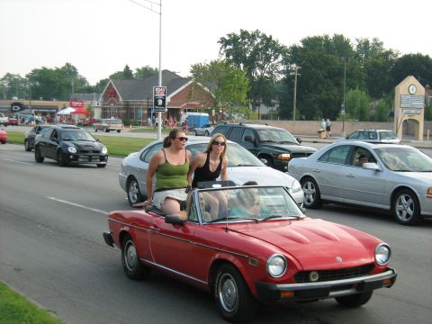 1988 Mercury Grand Marquis - People enjoy a ride in a red convertible during a car event, with other vehicles on the road.