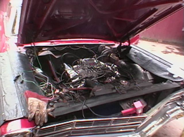 Open hood of maroon car showing engine bay with wiring, hand resting on front edge.