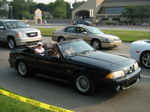 1988 Mercury Grand Marquis - A black convertible Mustang with a '5.0' badge cruises down the road, seen from the front-right angle.