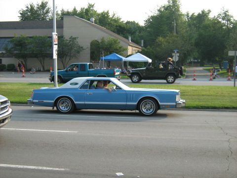 1988 Mercury Grand Marquis - A blue 1977 Mercury Cougar coupe with a white vinyl roof cruises down the road, with other vehicles and tents visible in the background.