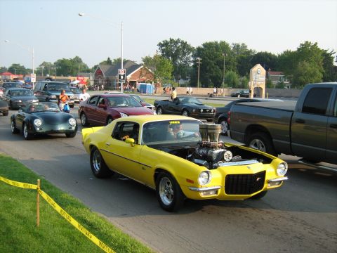 1988 Mercury Grand Marquis - A bright yellow muscle car with its hood open, revealing a large supercharger, cruises in traffic during a car show.