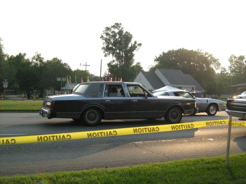 1988 Mercury Grand Marquis - A dark sedan is parked on the street with caution tape in front of it and other vehicles in the background.