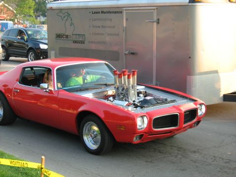1988 Mercury Grand Marquis - A red muscle car with its hood open, revealing a powerful engine, drives past a utility trailer.
