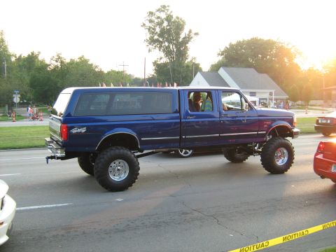 1988 Mercury Grand Marquis - A lifted blue crew cab pickup truck with a camper shell is seen driving on a street.