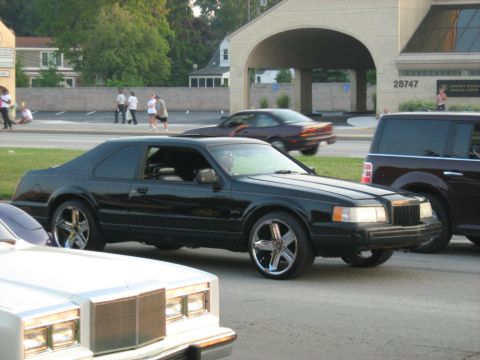 1988 Mercury Grand Marquis - A black coupe with custom wheels is parked on the street, with other vehicles and pedestrians in the background.