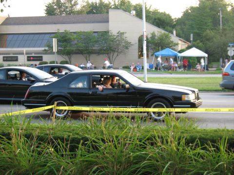 1988 Mercury Grand Marquis - A black coupe drives by a gathering of people under tents, with caution tape in the foreground.