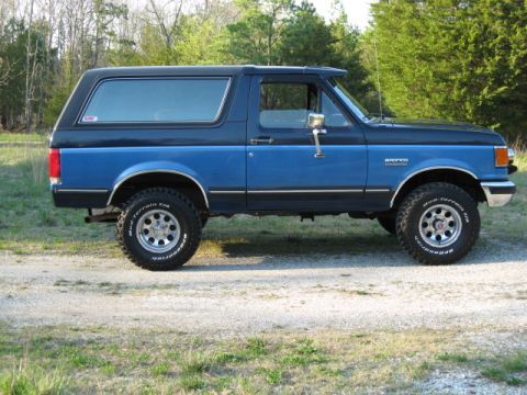 1990 Ford Bronco - A two-tone blue and black Ford Bronco is viewed from its side profile on a gravel road with trees in the background.