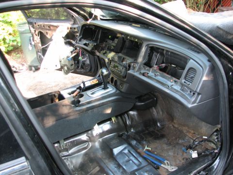 1999 Ford Crown Victoria - Interior teardown of a black sedan photographed through the open passenger door, with the grey dash pad in place but the instrument cluster…