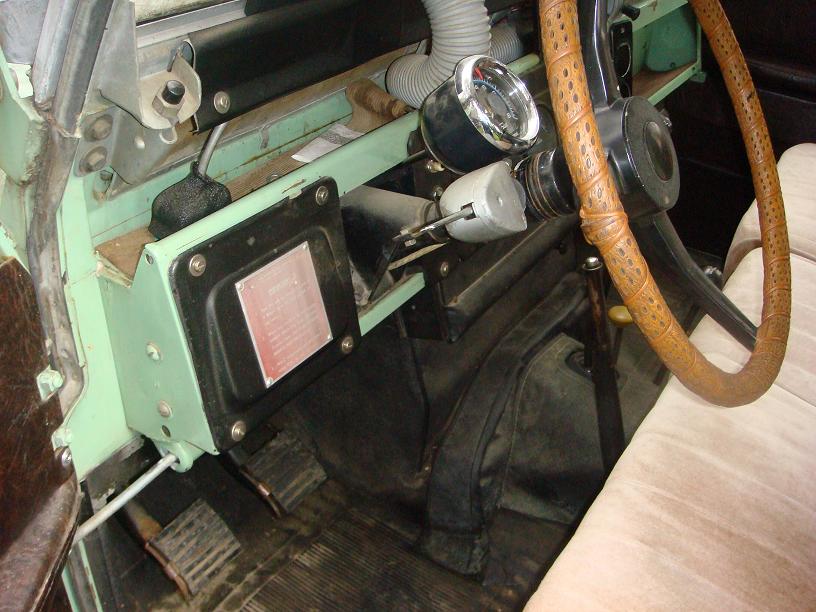 Vehicle interior with light green dashboard, brown steering wheel, and floor pedals. A metal plate with red text is visible