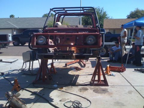 Suzuki Samurai - A red Suzuki Samurai stripped to its bare tub sits high on jack stands in a driveway, fitted with a tubular roll cage and round headlights…
