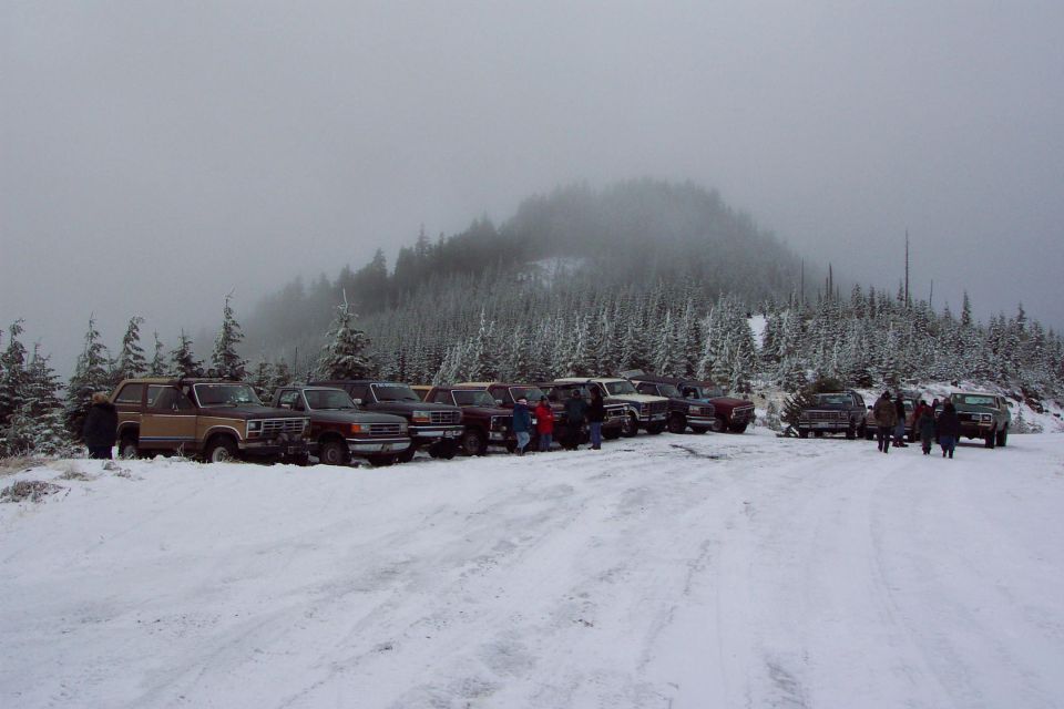 Row of Ford trucks and Broncos parked on a snowy mountain road with people standing nearby in fog.