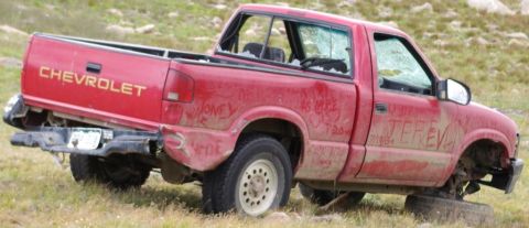 1995 Ford Bronco - A damaged red Chevrolet pickup truck sits in a grassy field with visible graffiti and missing parts.