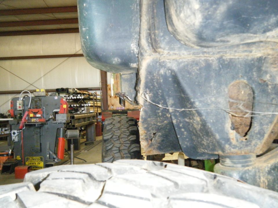 Close-up of a dark-colored vehicle frame and a large tire, with industrial machinery in the background