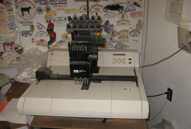 Embroidery machine with multiple needles and control panel, set against a wall covered in stickers and patches