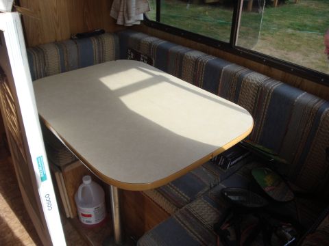 1988 Ford F-350 - The interior of a camper van shows a dinette table and striped upholstered seating, with a window overlooking a grassy area.