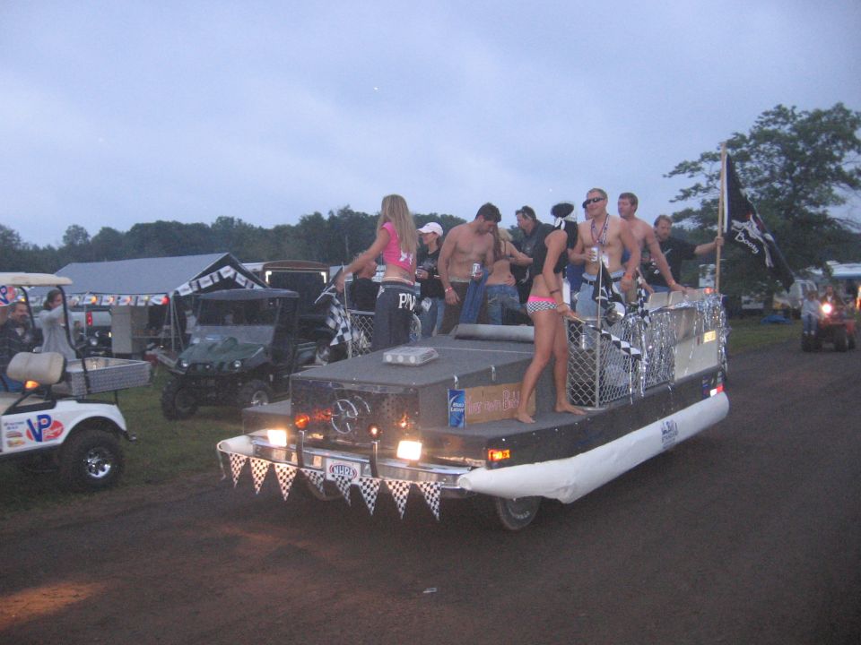 People riding a custom pontoon-boat parade float at a campground at dusk