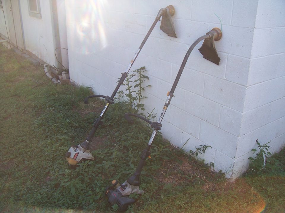 Two string trimmers leaning against a white brick wall, one on the left and one on the right, with grass and weeds around them