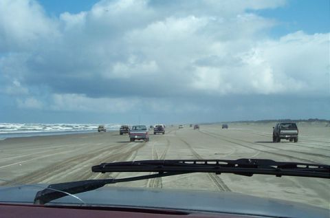 1989 Ford F-150 - Multiple trucks drive along a wide, sandy beach with the ocean on the left and dunes on the right under a cloudy sky.