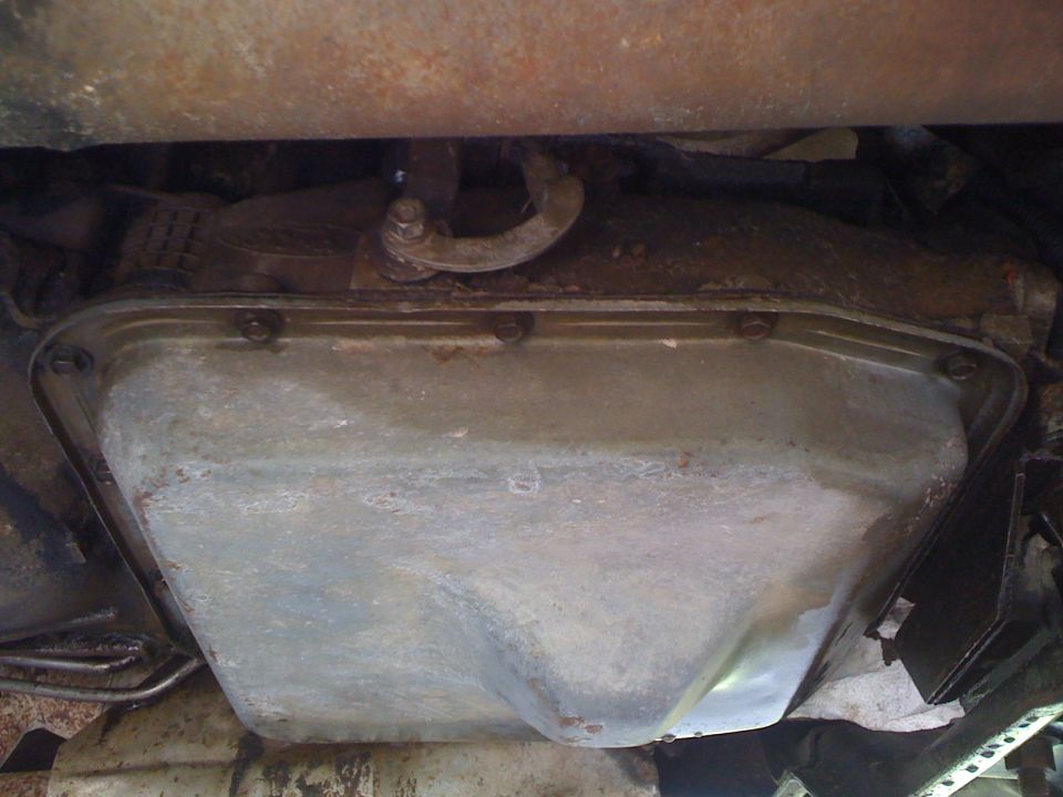 Underside view of a dirty stamped-steel automatic transmission pan with bolts and shift linkage above
