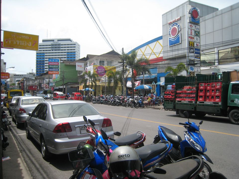 Busy street scene with parked cars, motorbikes and shops with Thai signage