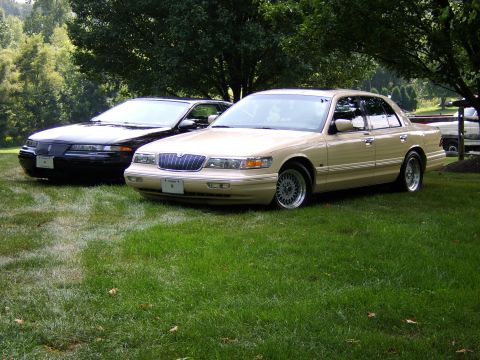 1996 Mercury Grand Marquis - A tan Mercury Grand Marquis and a black Ford Crown Victoria are parked on a grassy lawn under trees.
