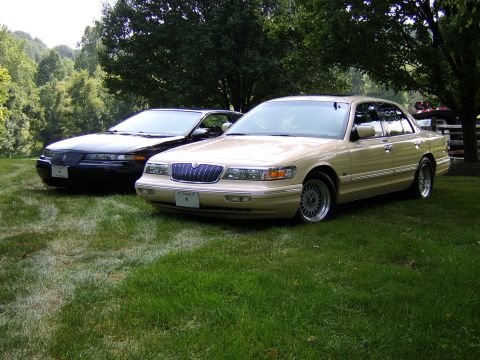 1996 Mercury Grand Marquis - Two sedans are parked on a grassy lawn under trees, with a wooden fence visible in the background.