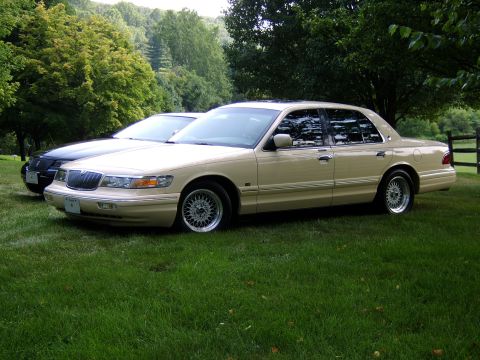 1996 Mercury Grand Marquis - Two sedans are parked on a green lawn with trees and a wooden fence in the background.