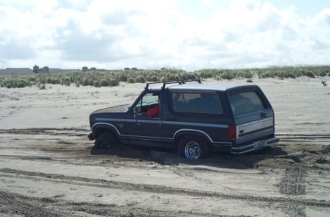 1989 Ford F-150 - A Ford Bronco is stuck in the sand near dunes with tire tracks visible.