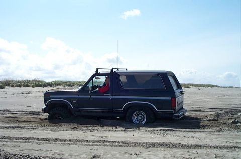 1989 Ford F-150 - A dark blue Ford Bronco is stuck in the sand on a beach, with a driver visible behind the wheel.