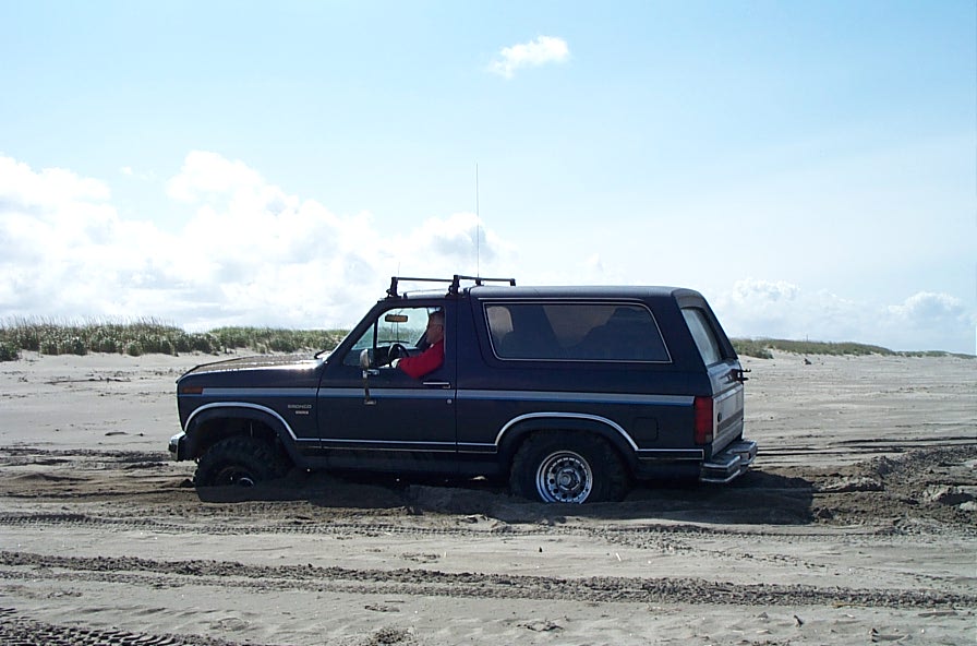 Dark blue 1980s Ford Bronco stuck axle-deep in beach sand, side view with dunes behind