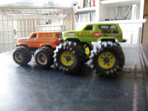 1986 Ford Bronco - Two toy monster trucks are parked next to each other on a dark surface, with a white cage in the background.