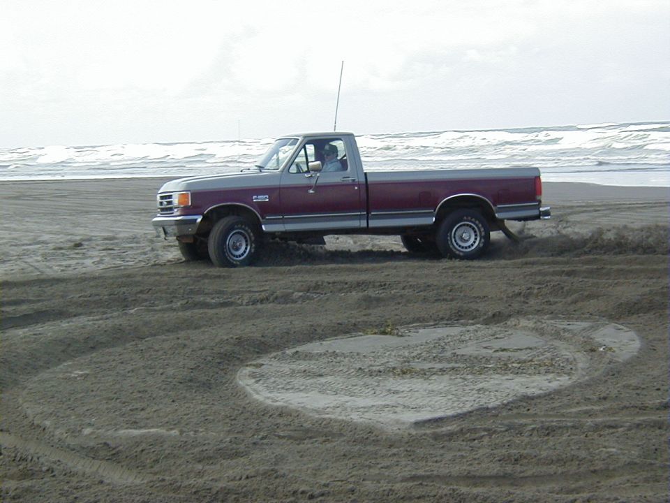 Maroon and silver two-tone Ford F-150 4x4 cutting circles in beach sand near the surf