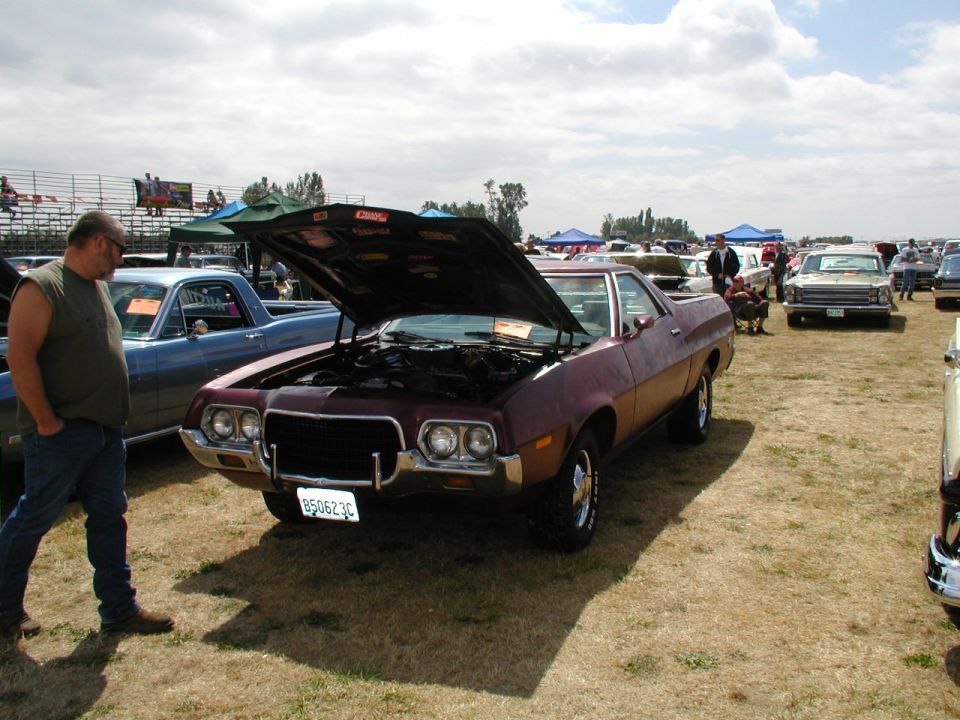 Maroon 1972 Ford Ranchero with hood open, parked at a car show next to a blue pickup
