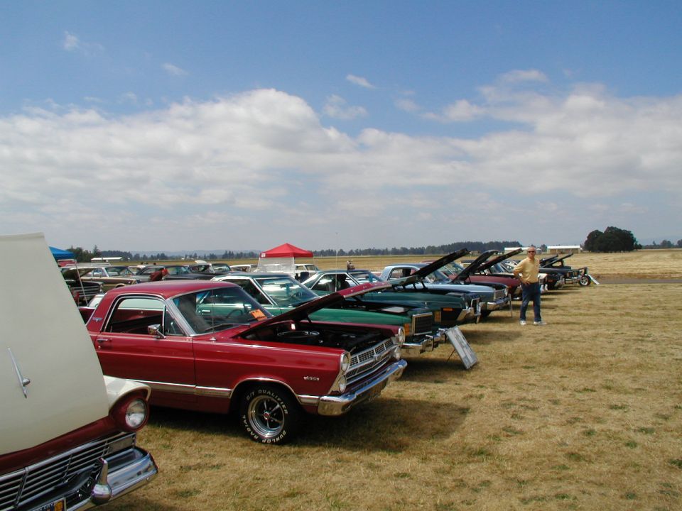 1967 Ford Ranchero in a line of cars with hoods up at a show
