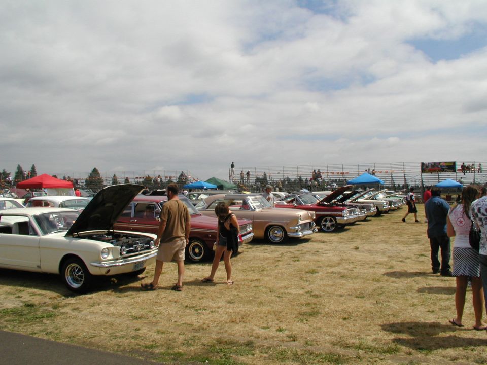 White 1965 Ford Mustang coupe with hood open, parked on dry grass at a car show