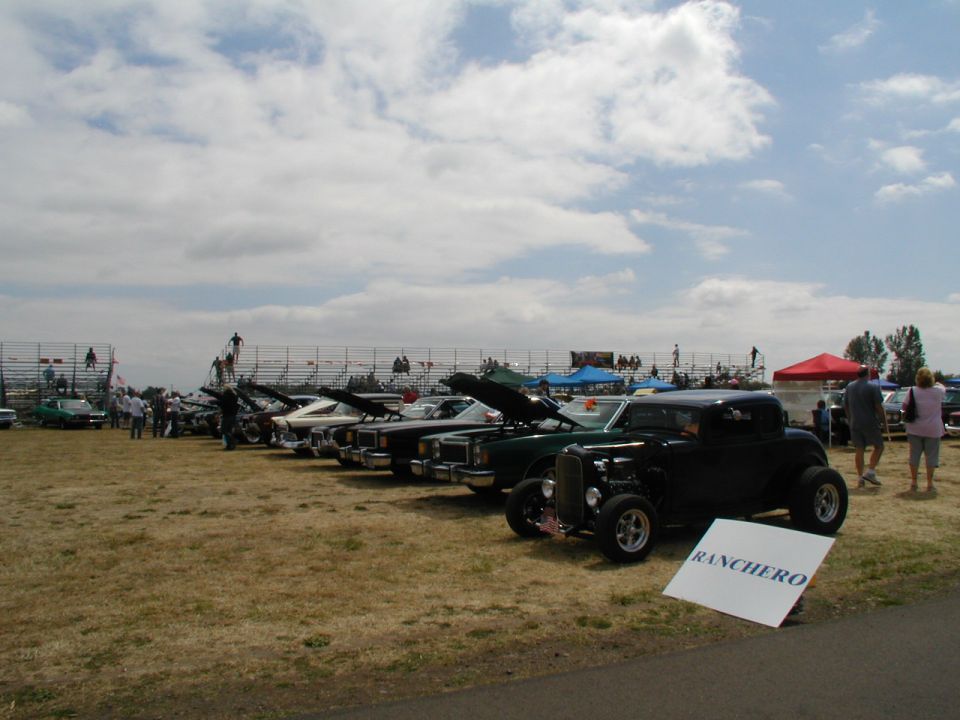 Black hot rod with exposed engine and custom wheels, parked on grass with a sign that reads RANCHERO